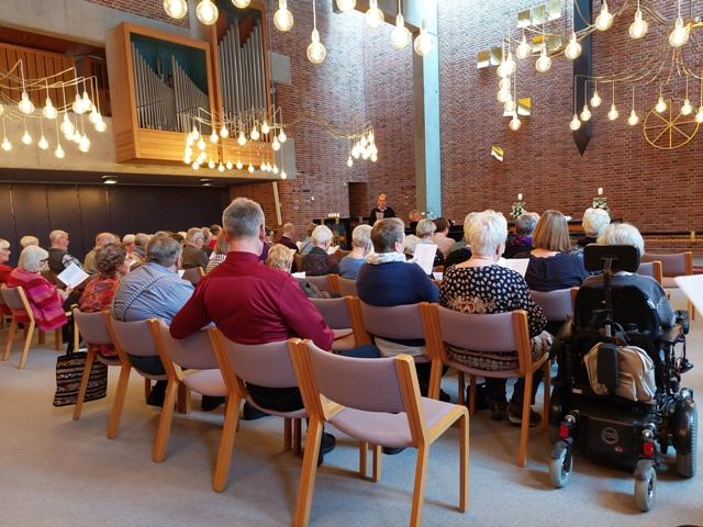 En gruppe mennesker sidder i en kirke og synger, mens de holder sanghæfter. Der er en orgel og smukke lamper i rummet.