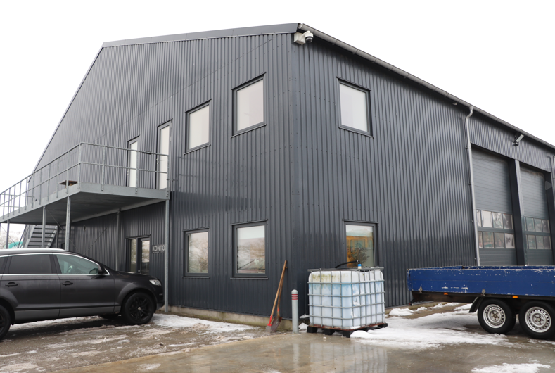 En moderne, grå bygning med store vinduer, en trappe og en parkeret bil foran.