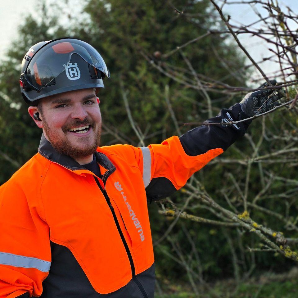 En glad mand i orange sikkerhedstøj og hjelm, der beskærer grene i en have.