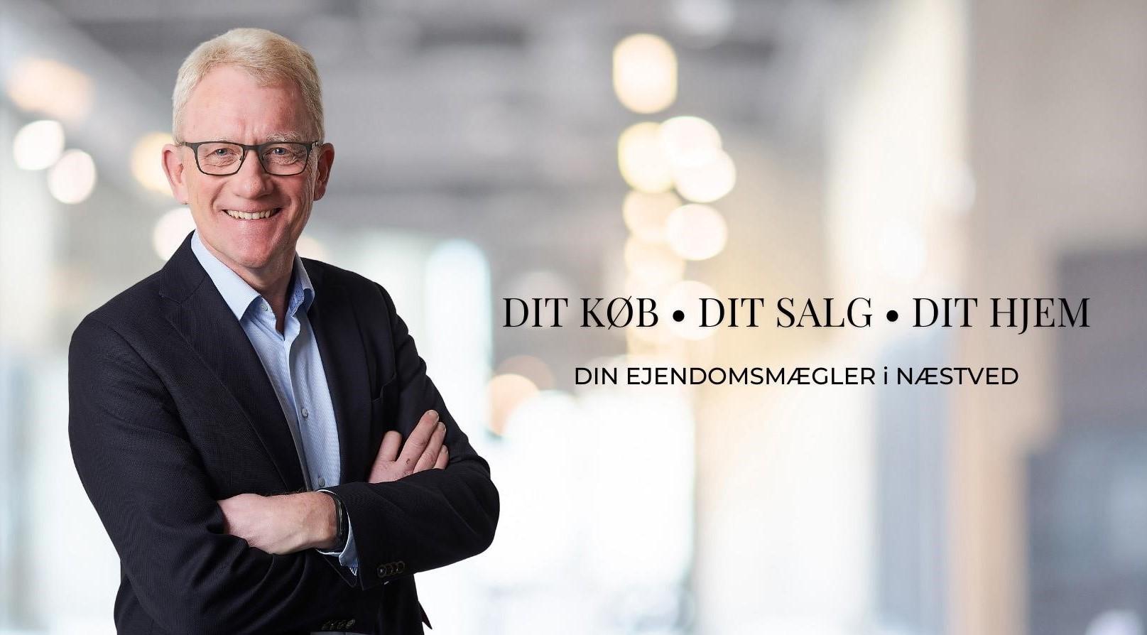En smilende mand i jakkesæt står med armene over kors i et lyst kontormiljø. Tekst om ejendomshandler i baggrunden.