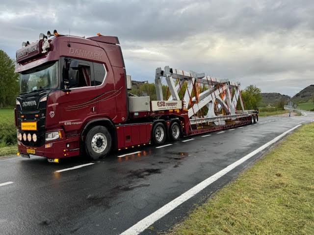 En stor rød lastbil transporterer en stor metalstruktur på en vej under en overskyet himmel.