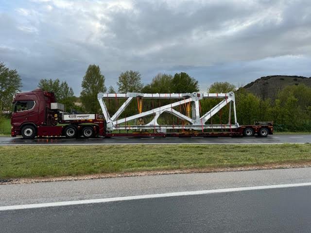 En stor lastbil transporterer en hvid stålramme langs en vej med grønne træer i baggrunden.