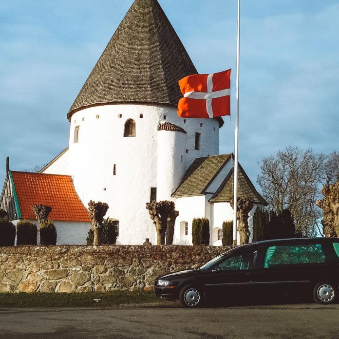 En hvid rund kirke med spidst tag, en dansk flag og en sort bil parkeret foran.