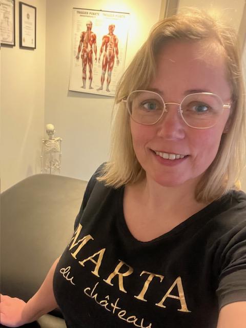 Kvinde med briller iført en sort t-shirt med teksten "MARTA du château" i et behandlingsrum.