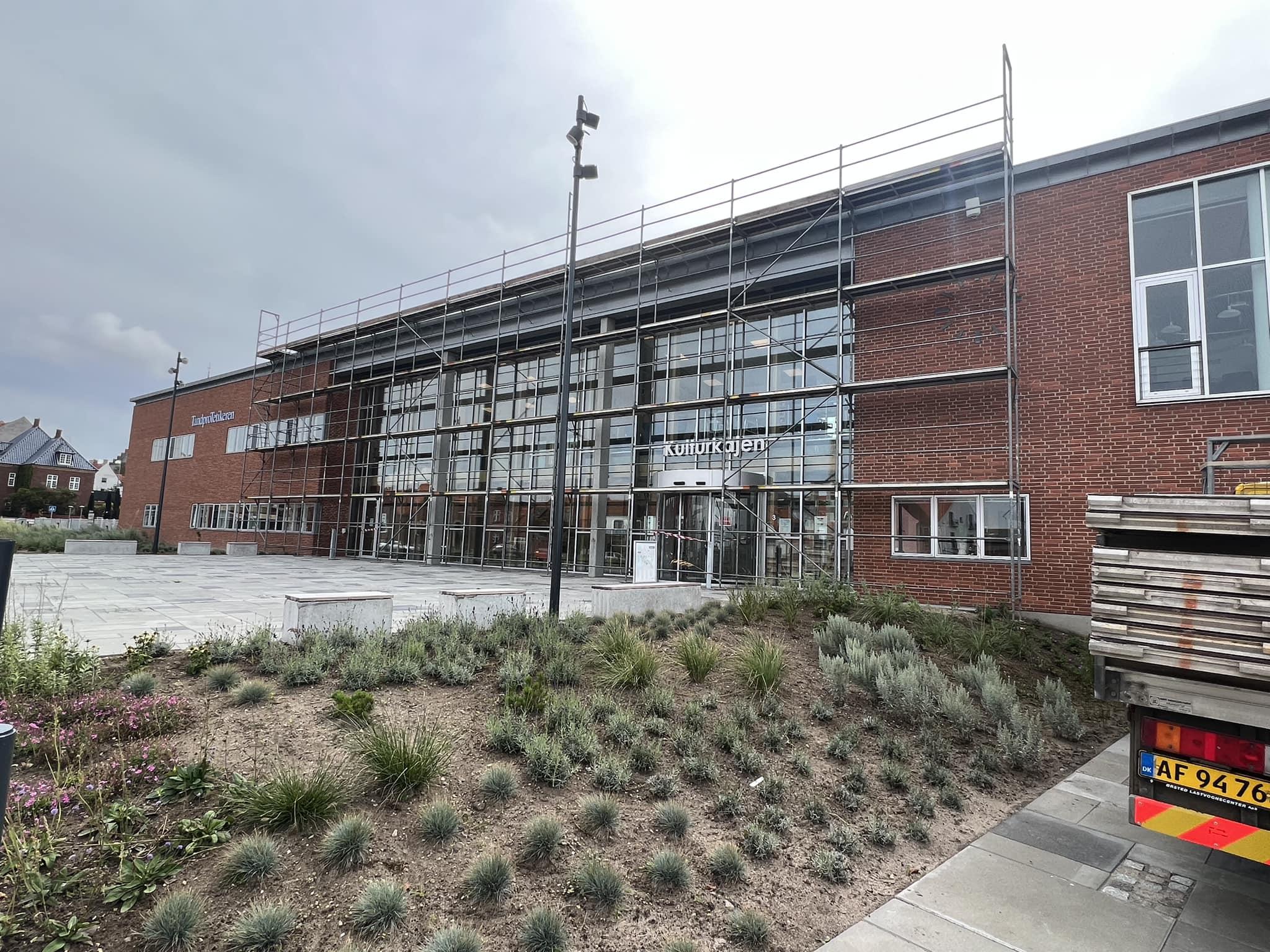 Bygning med glasfacade og stillads, omgivet af beplantning og en parkeringsplads.
