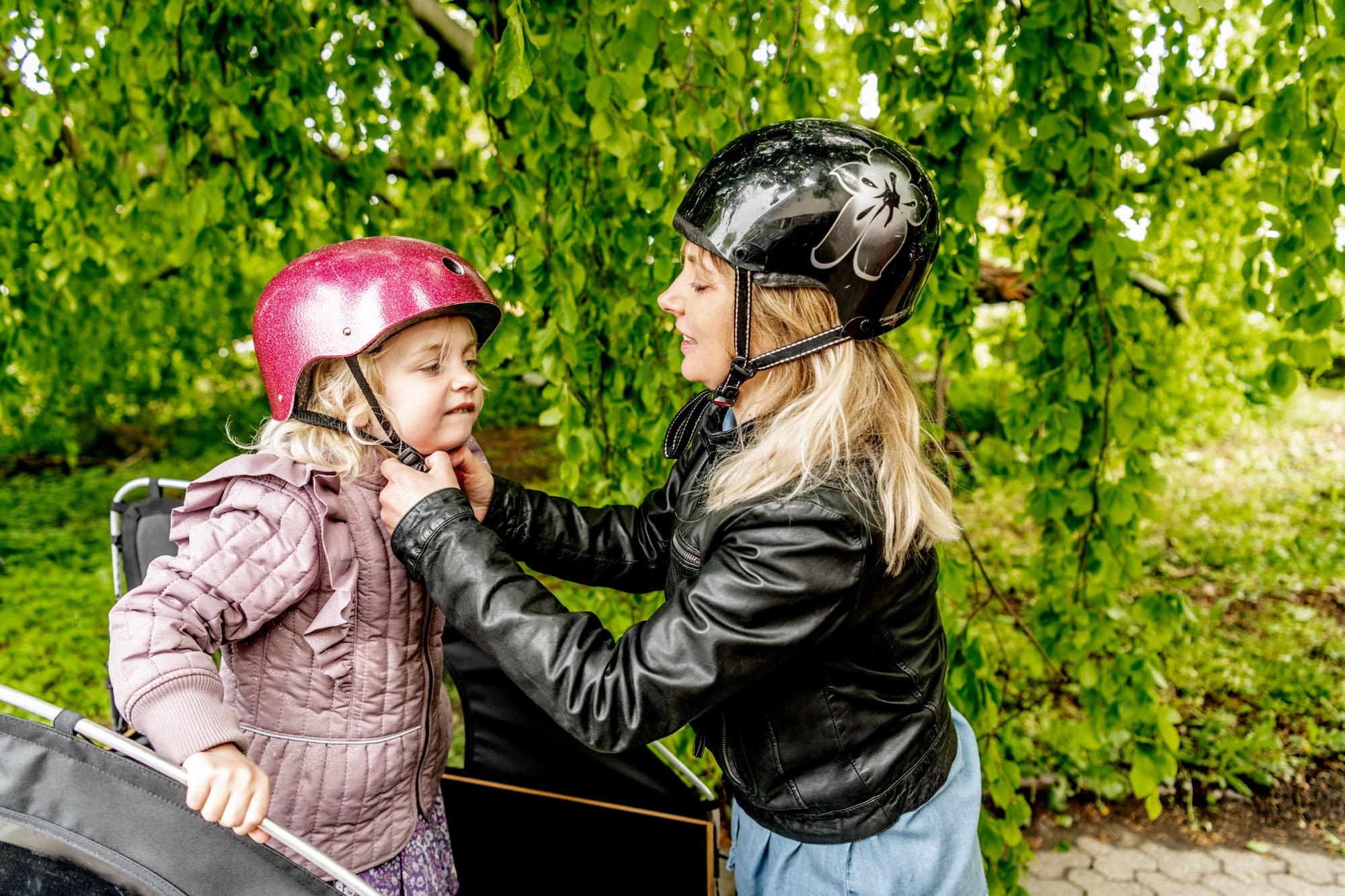 To piger iført cykelhjelme står under et træ med grønne blade, mens den ene hjælper den anden med hjelmen.