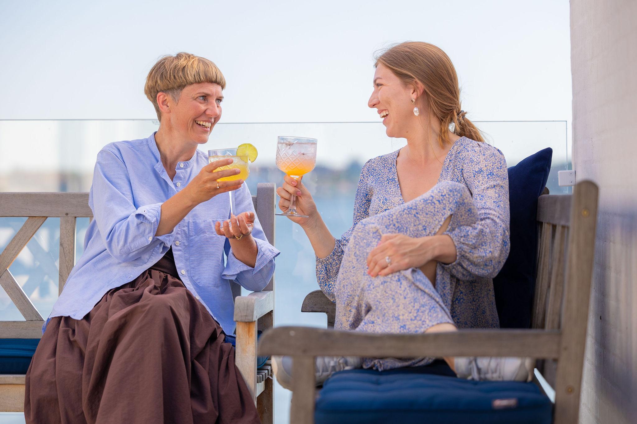 To kvinder sidder på en bænk og skåler med farverige drinks på en solrig terrasse.