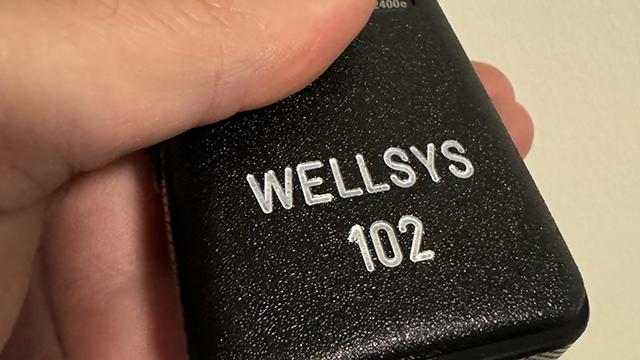 En hånd holder en sort enhed med teksten "WELLSYS 102" på siden.