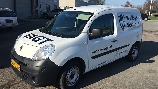 Hvid Renault varevogn med "Wellys Security" logo og hjemmeside på siden, parkeret på en grusvej.