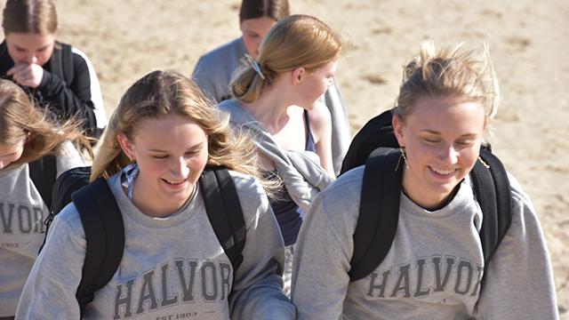 To unge piger iført grå sweatshirts med rygsække, der smiler og går op ad en sandbakke.