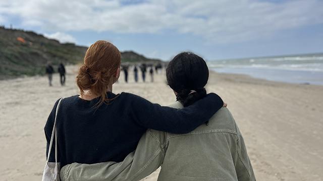 To personer går sammen på stranden med armene om hinanden, mens de ser ud mod havet og en gruppe mennesker i baggrunden.