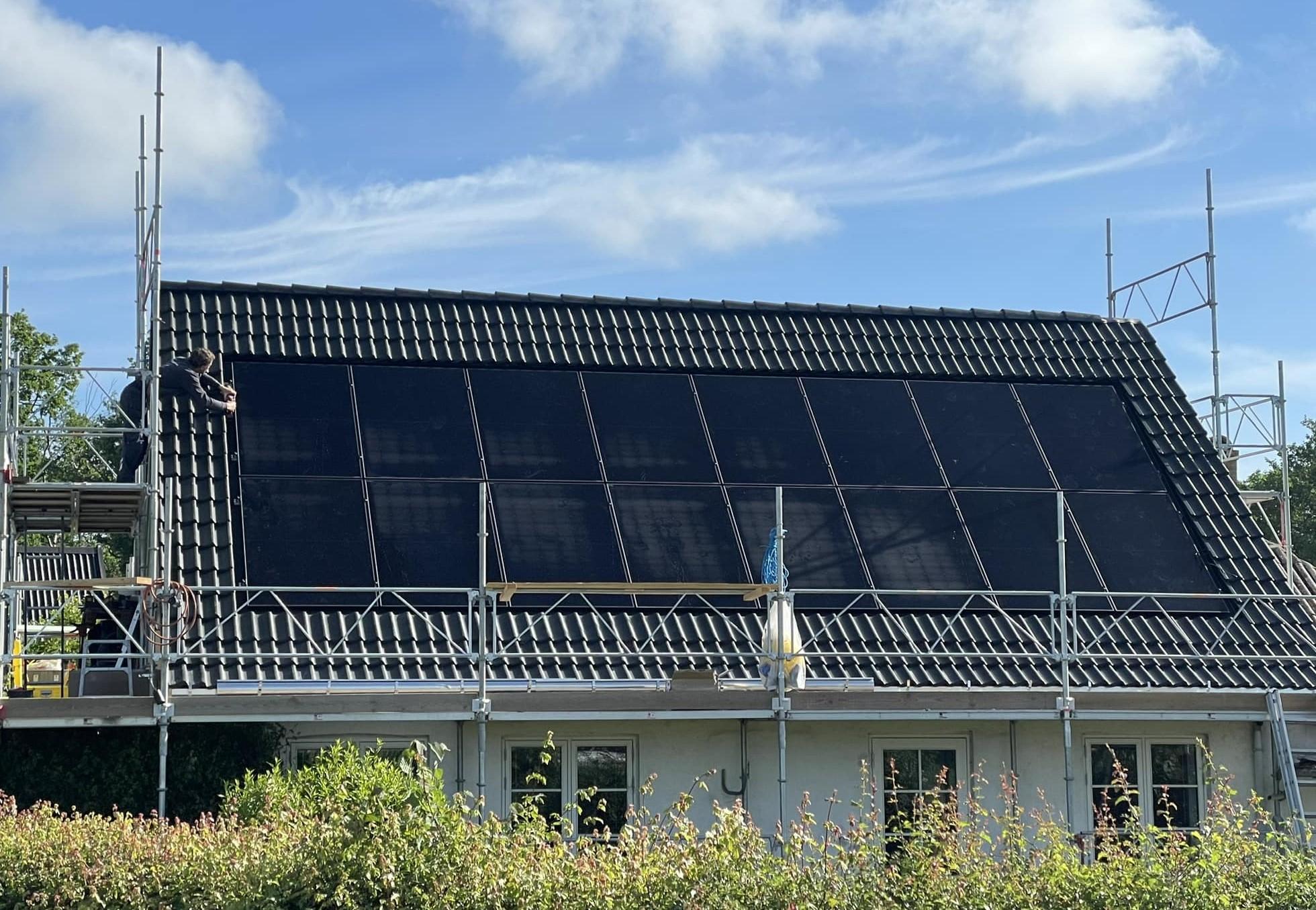 Hus med solpaneler på taget, omgivet af stilladser og grønne planter i forgrunden.