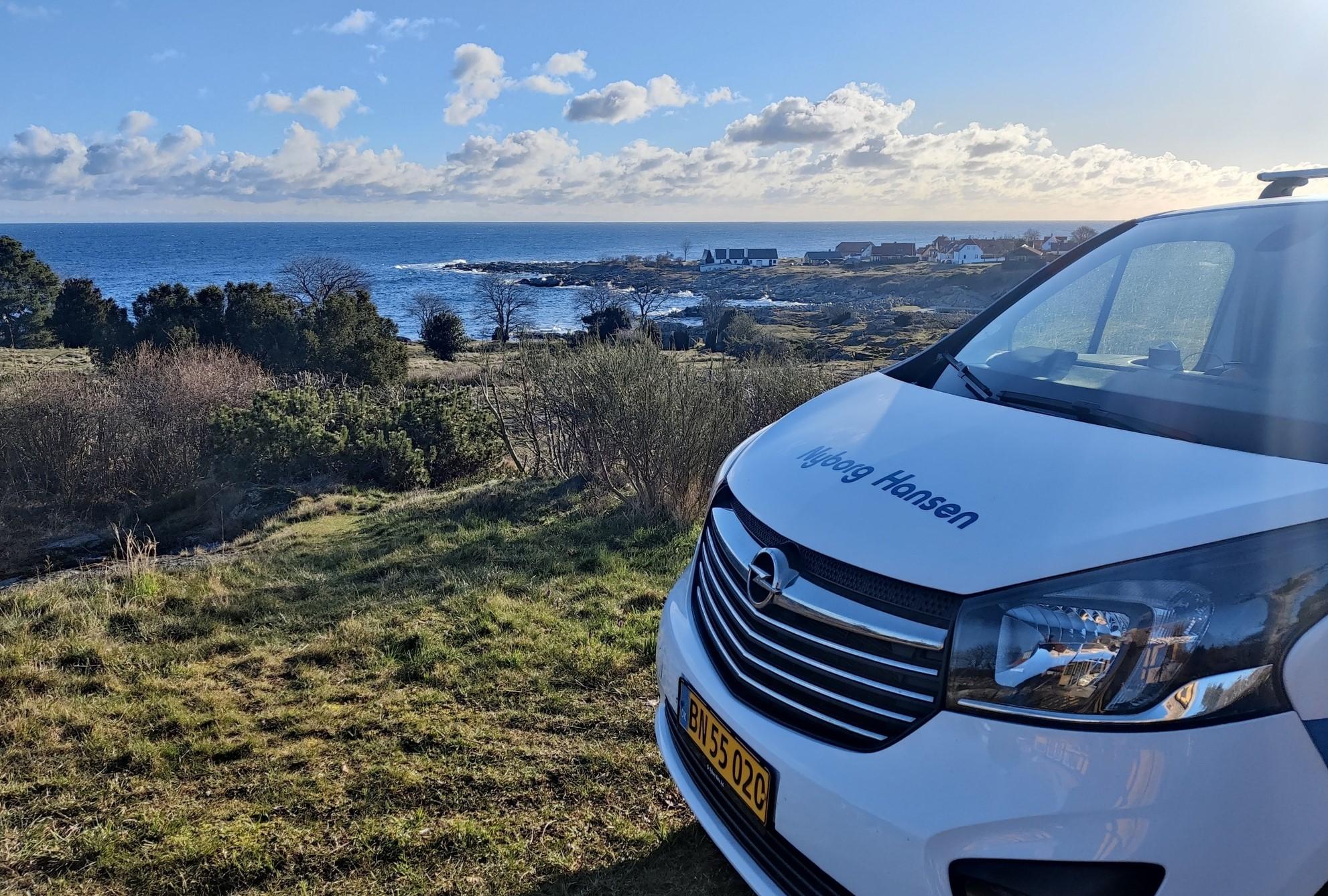 Udsigt over havet med en hvid bil i forgrunden og en kystby i baggrunden.