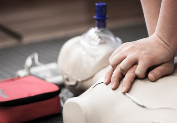 Hånd, der udfører hjertemassage på en førstehjælpsdukke med en hjertestarter ved siden af.