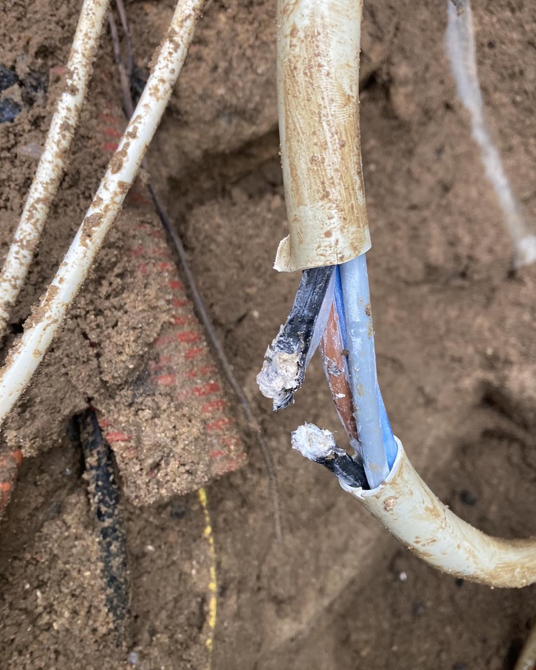 Skadet elektrisk kabel med synlige ledninger i jorden, omgivet af jord og andre kabler.