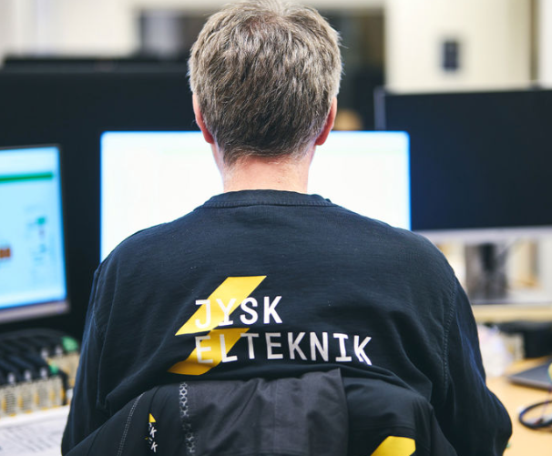 En person med ryggen til, iført en sort trøje med teksten "JYSK ELTEKNIK", sidder foran computere.