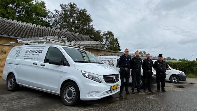 Fem mænd i uniform står foran to hvide varevogne med logoer, på en grå dag med skyer.
