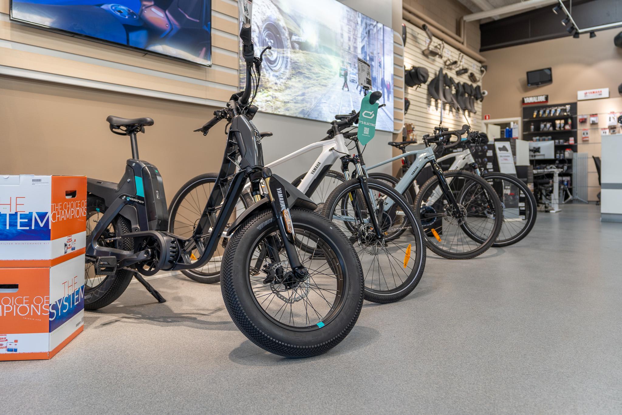 Flere elcykler udstillet i en moderne cykelbutik med tilbehør og skærme i baggrunden.