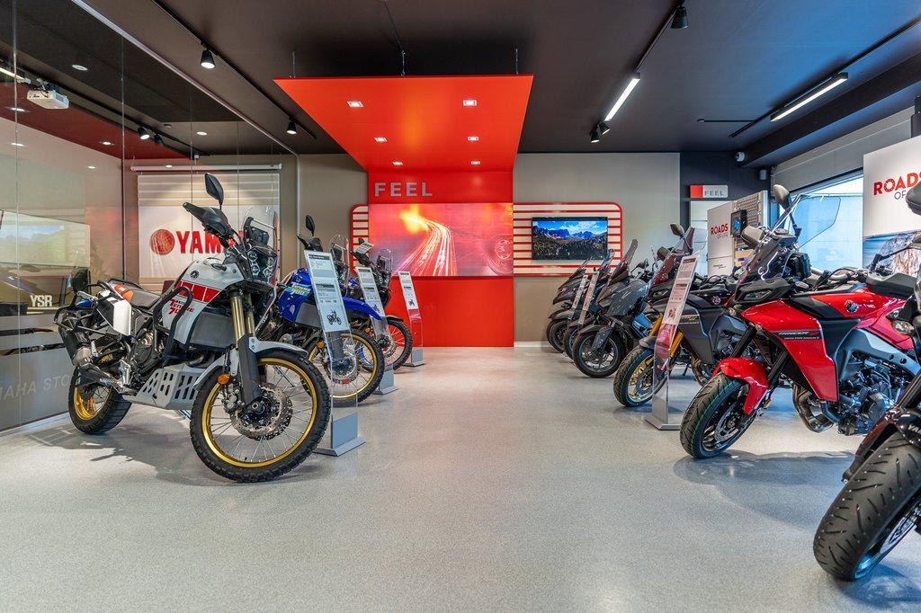 En moderne motorcykelbutik med flere motorcykler udstillet, herunder Yamaha-modeller og en stor skærm.