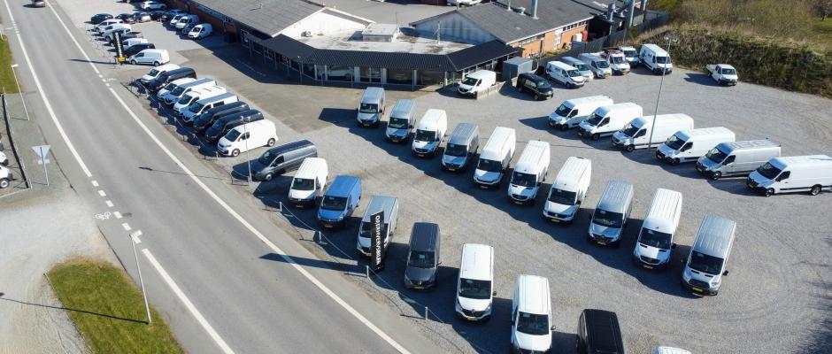 Oversigt over parkerede varevogne og biler på en grusplads ved en bygning langs vejen.