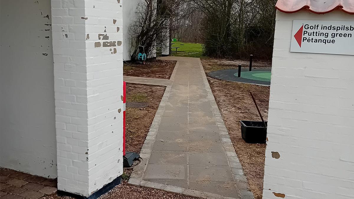 En grussti fører gennem en hvid mur med en golf putting green til venstre og grønne områder i baggrunden.