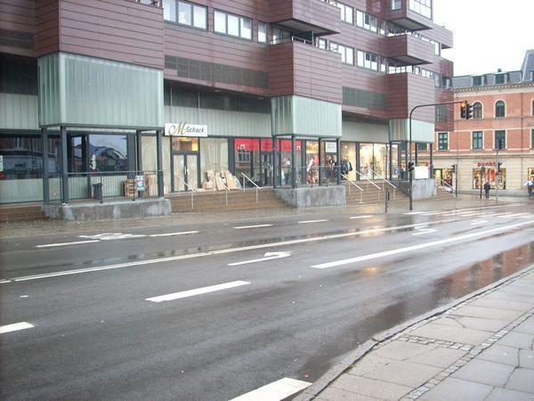 En gade med butikker i stueetagen af en moderne bygning, regnvejr og reflekterende vand på vejen.