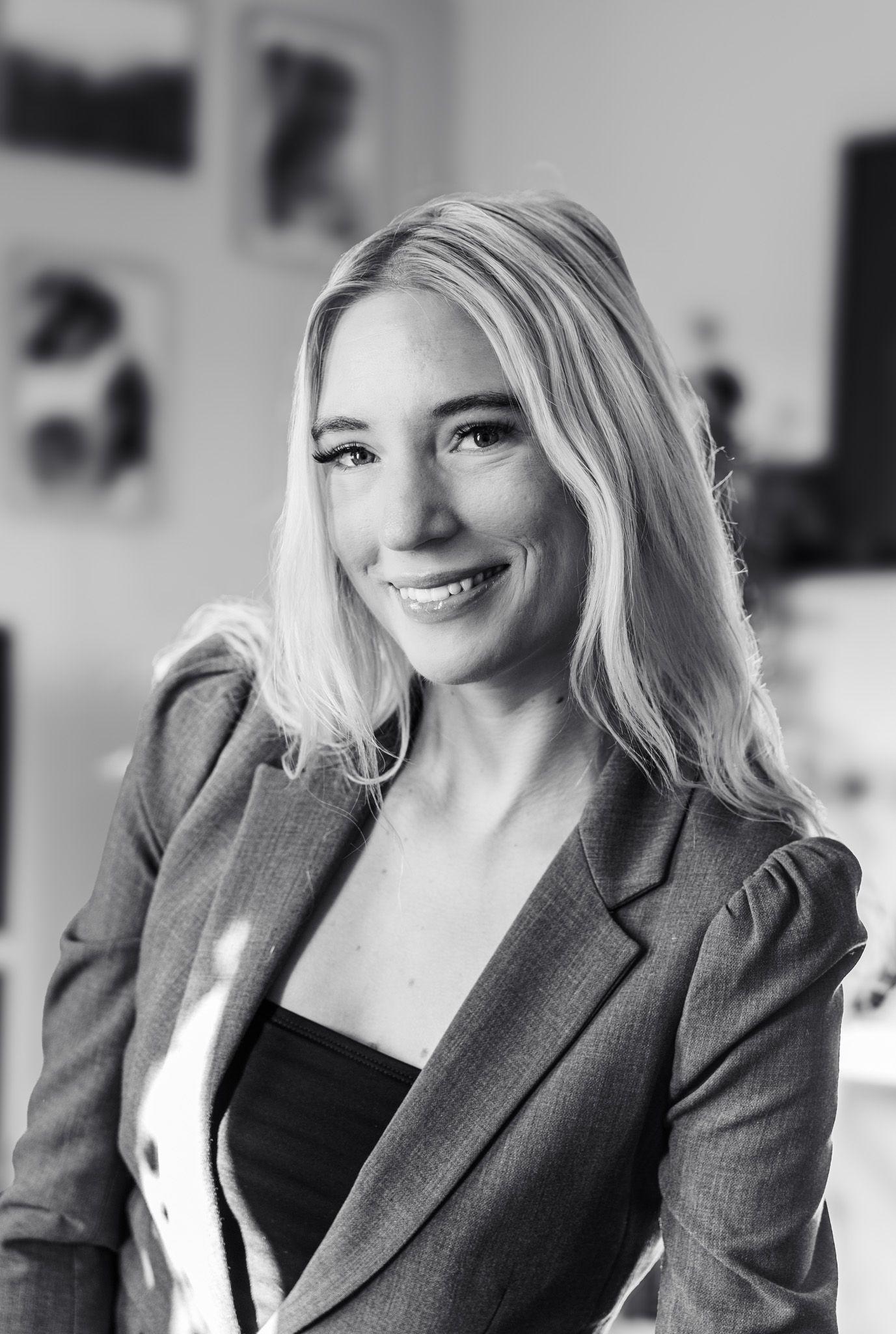 En smilende kvinde med langt, lyst hår iført en grå blazer, står i et lyst rum med billeder i baggrunden.