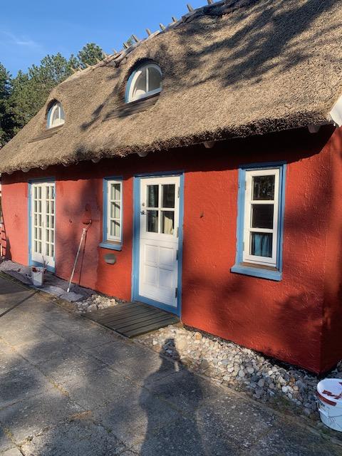 Et rødt hus med stråtag og blå vinduer, beliggende på en grusvej under en klar blå himmel.