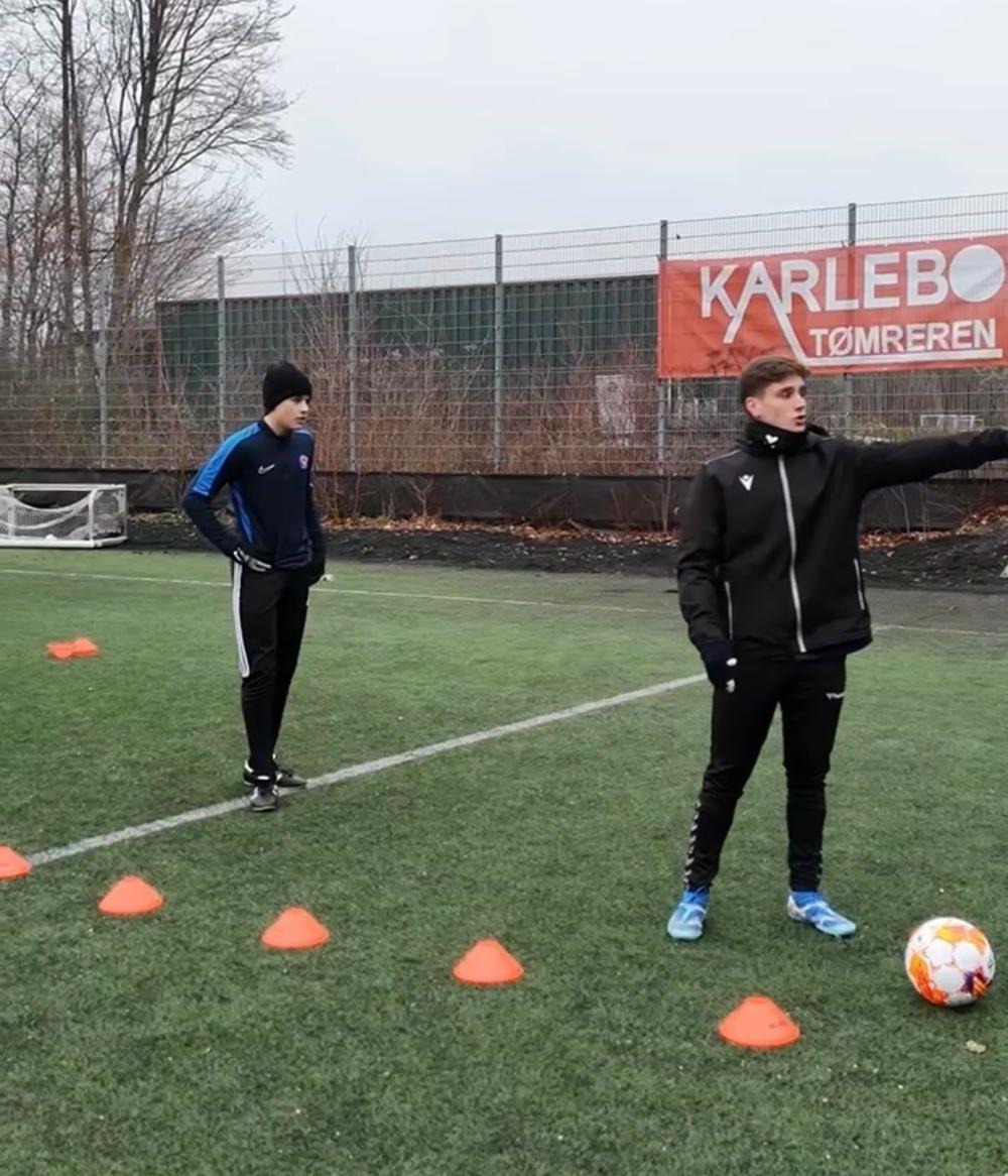Træner peger mod en gruppe orange kegler på en fodboldbane, mens en spiller står i baggrunden.