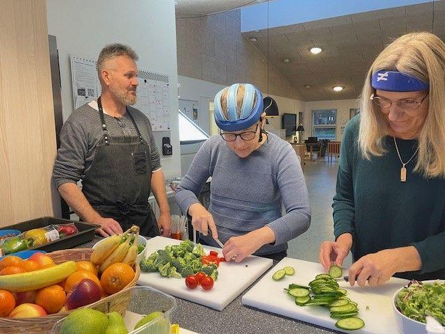 Tre personer laver mad i et køkken, hvor de skærer grøntsager som agurker og broccoli.