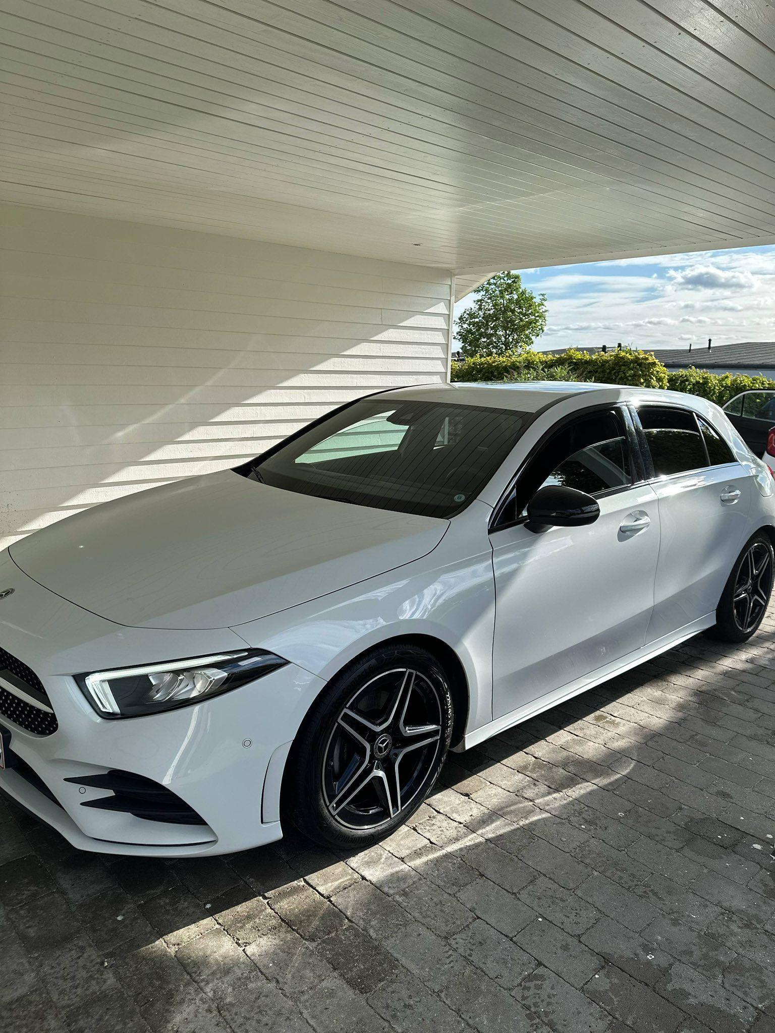 En hvid Mercedes-Benz A-klasse parkeret under en carport med sollys og grønne planter i baggrunden.