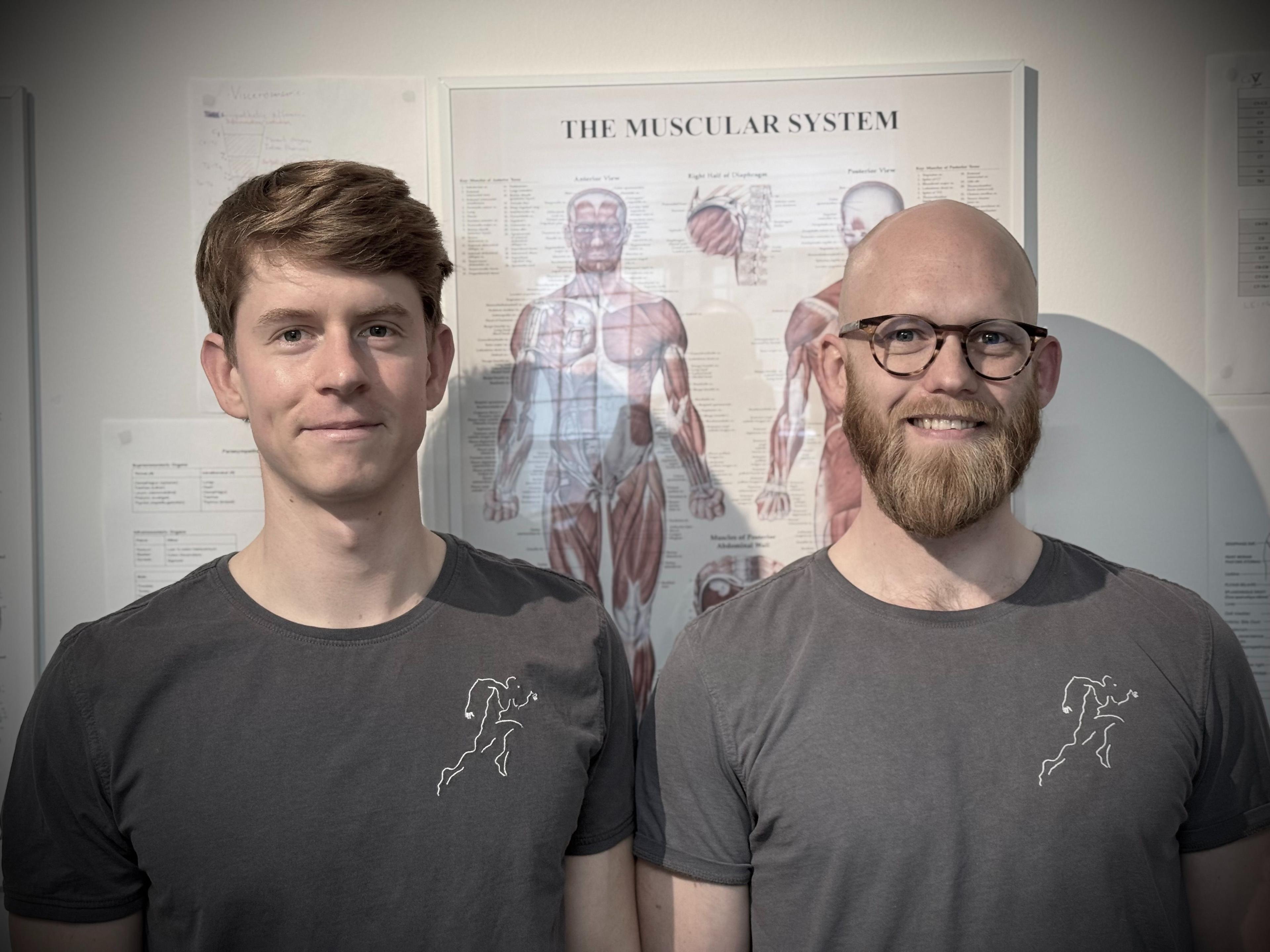 To personer står foran en plakat med det muskulære system, iført grå t-shirts.