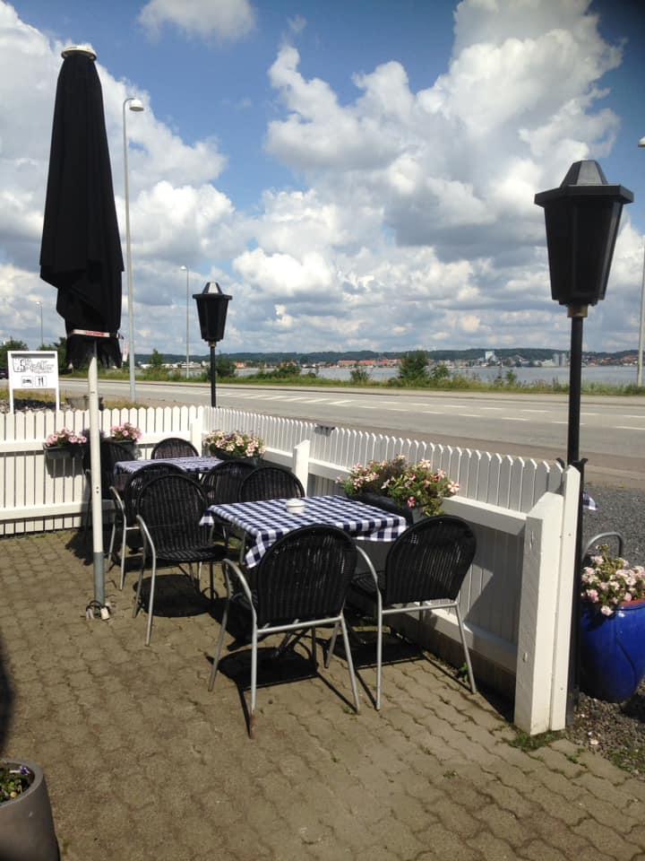Udeservering med sorte stole og ternede borde, omgivet af blomster og en smuk himmel med skyer.