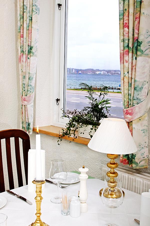 Et elegant spisebord med stearinlys, servietter og en lampe, med udsigt til havet gennem et vindue.