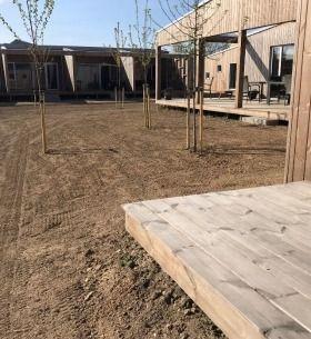 En nyanlagt gård med træterrasse og unge træer, omgivet af bygninger i træ under en klar blå himmel.
