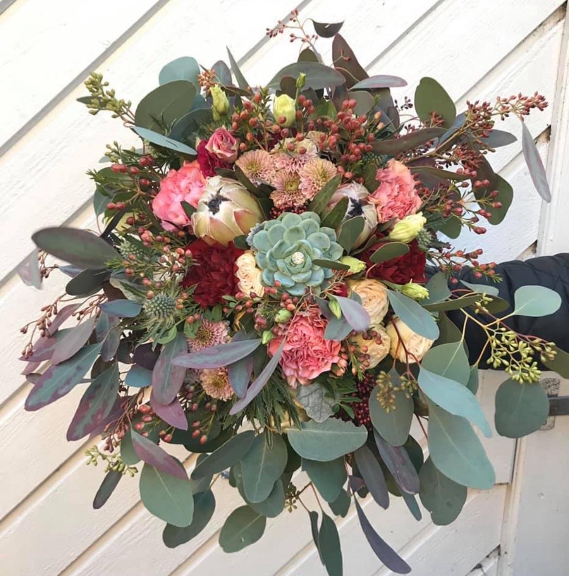 En farverig buket med roser, nelliker, succulenter og eucalyptus, præsenteret foran en hvid væg.