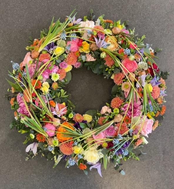 En farverig blomsterkrans med roser, nelliker og grønne blade på en grå overflade.