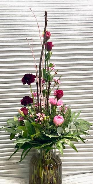 En smuk buket blomster med røde og lyserøde blomster, grønne blade og dekorative grene i en glasvase.