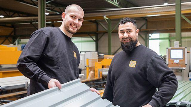 To mænd i en fabrik, der holder en metalplade, iført sorte trøjer med logoer, smilende og arbejder sammen.