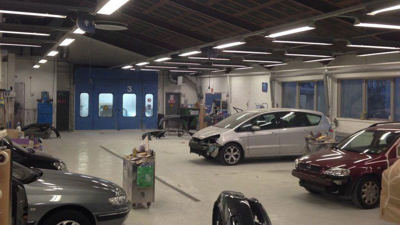 Et bilværksted med flere biler, malerudstyr og en blå garageport i baggrunden.