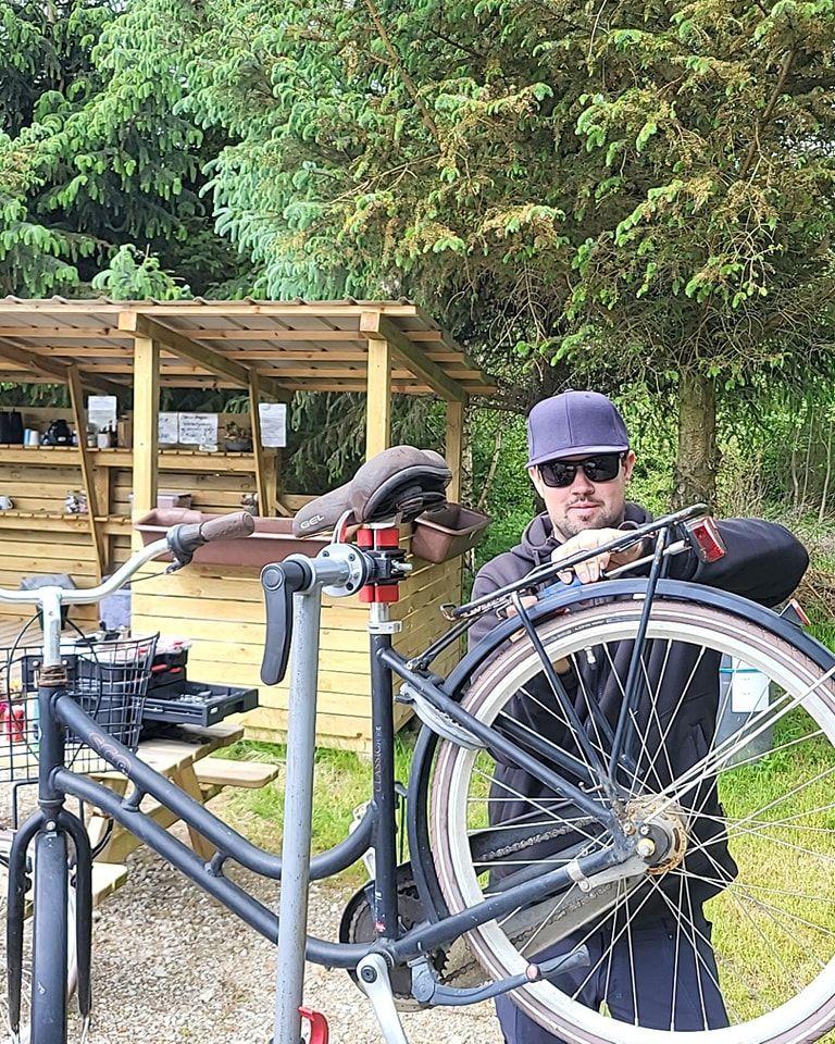En mand arbejder på en cykel ved en cykelværksted i naturen, iført solbriller og en kasket.