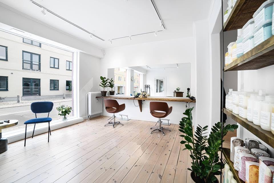 En moderne frisørsalon med to frisørstole, et spejl og planter i et lyst rum med store vinduer.