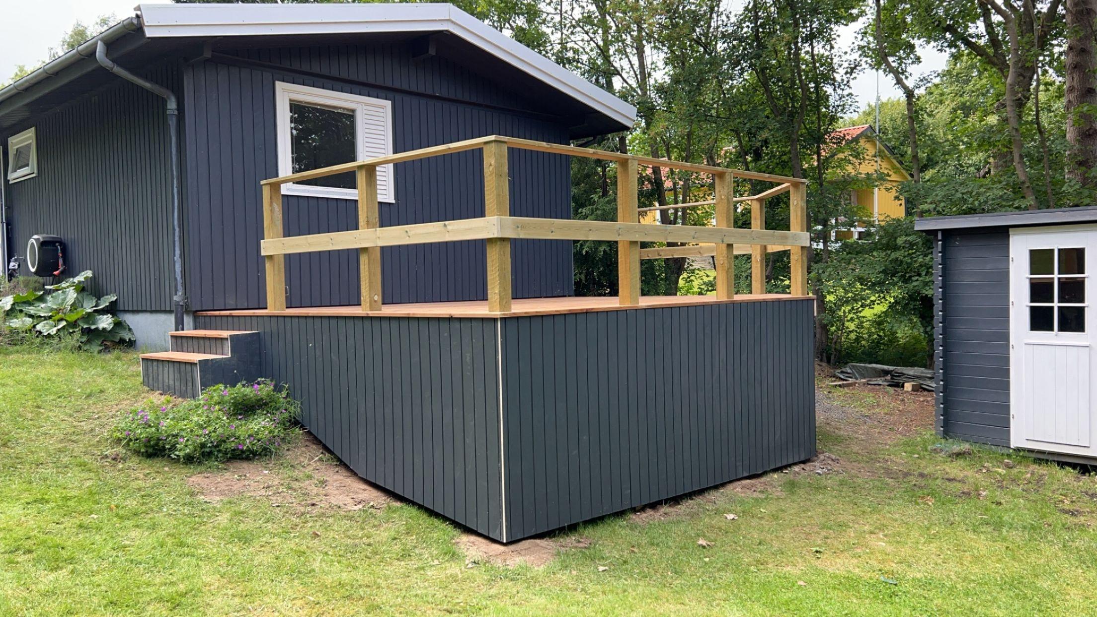 Et gråt hus med en træterrasse og gelænder, omgivet af grønne træer og græs.