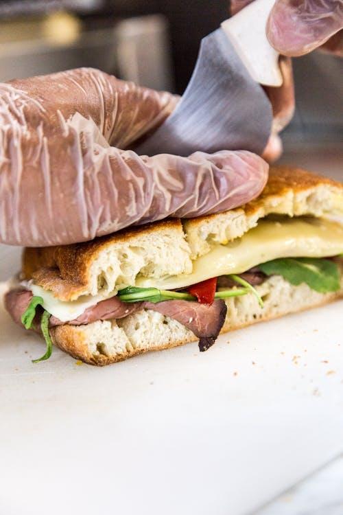 En hånd i plastikhandsker skærer en sandwich med skinke, ost, salat og peberfrugt på et skærebræt.