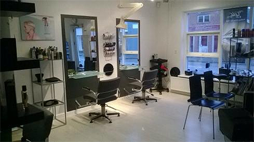 Frisørsalon med to frisørstole, spejle og produkter på hylder i et lyst rum.