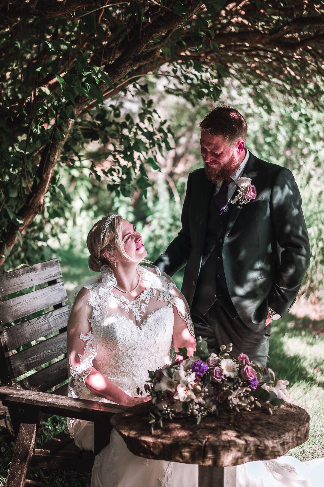 Brudepar sidder under en træbue, smilende og romantiske, med blomsterarrangement på bordet.
