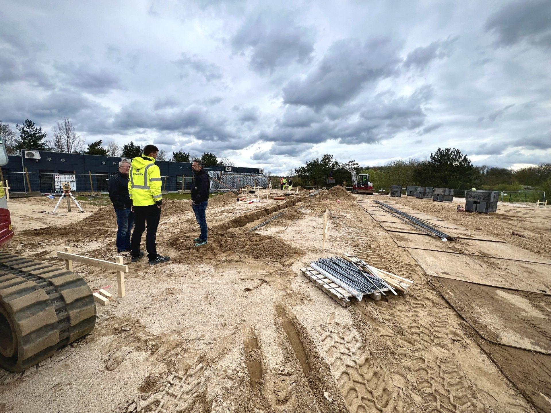 Byggeplads med sand, materialer og arbejdere iført sikkerhedstøj under en overskyet himmel.