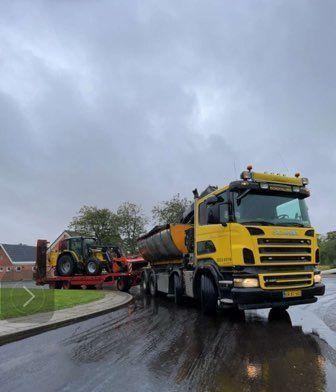 En gul lastbil med en stor tank og en traktor på en trailer kører på en våd vej under en grå himmel.