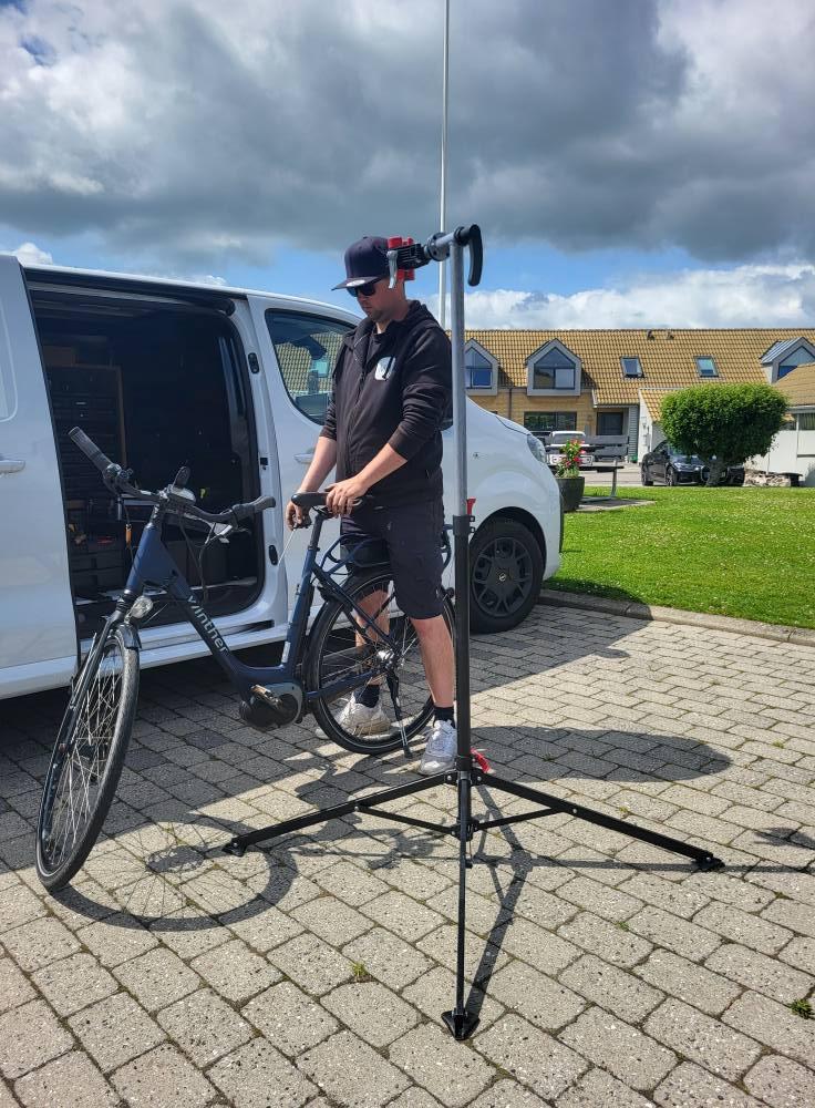 En mand står ved sin cykel på en cykelstand, mens han arbejder ved en hvid varevogn på en græsplæne.