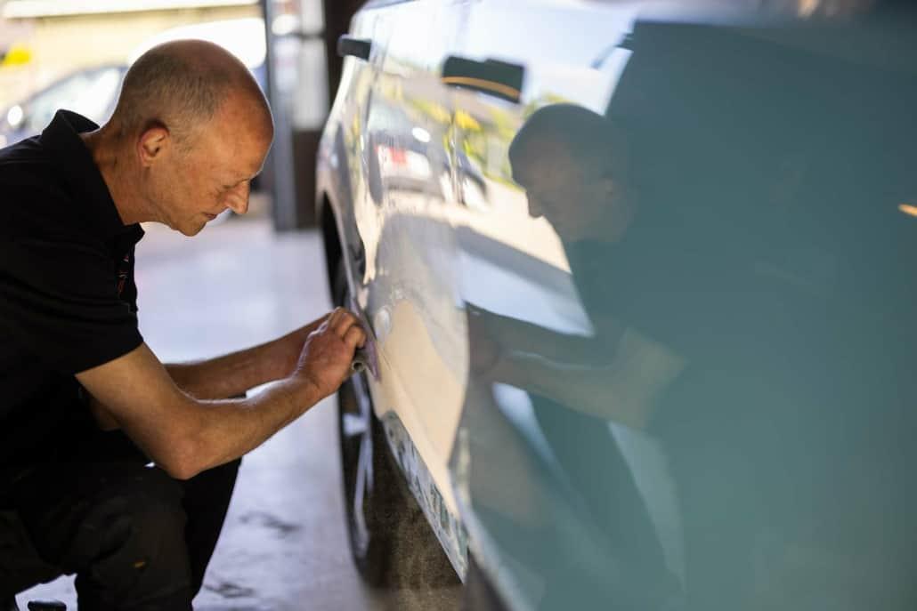 En mekaniker arbejder koncentreret på at reparere en bil i et værksted. Refleksionen af bilen ses tydeligt.